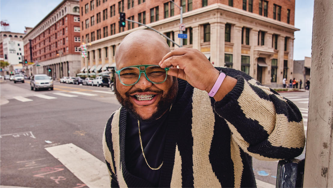 Homme souriant avec des lunettes vertes les ajustant dans une rue de la ville, portant un pull rayé.