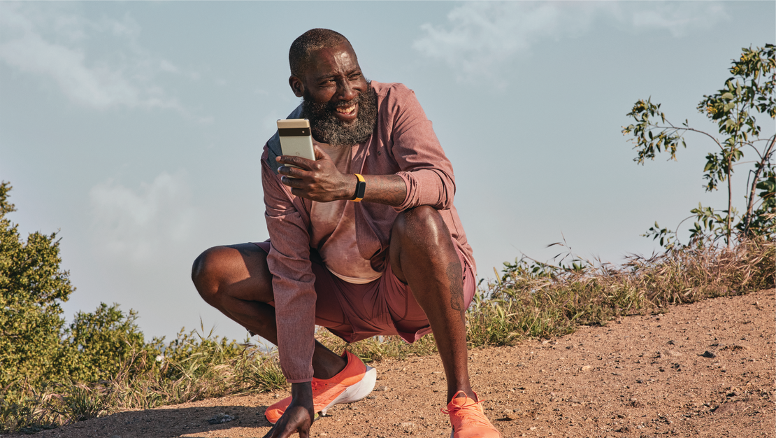 Homme en tenue de sport accroupi sur terre, tenant un téléphone, souriant, avec une montre au poignet.