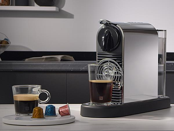 Eine silberne Kaffeemaschine mit zwei Kaffeegläsern. Drei Kaffeekapseln liegen auf einem kleinen Teller. Der Hintergrund ist eine Küche.
