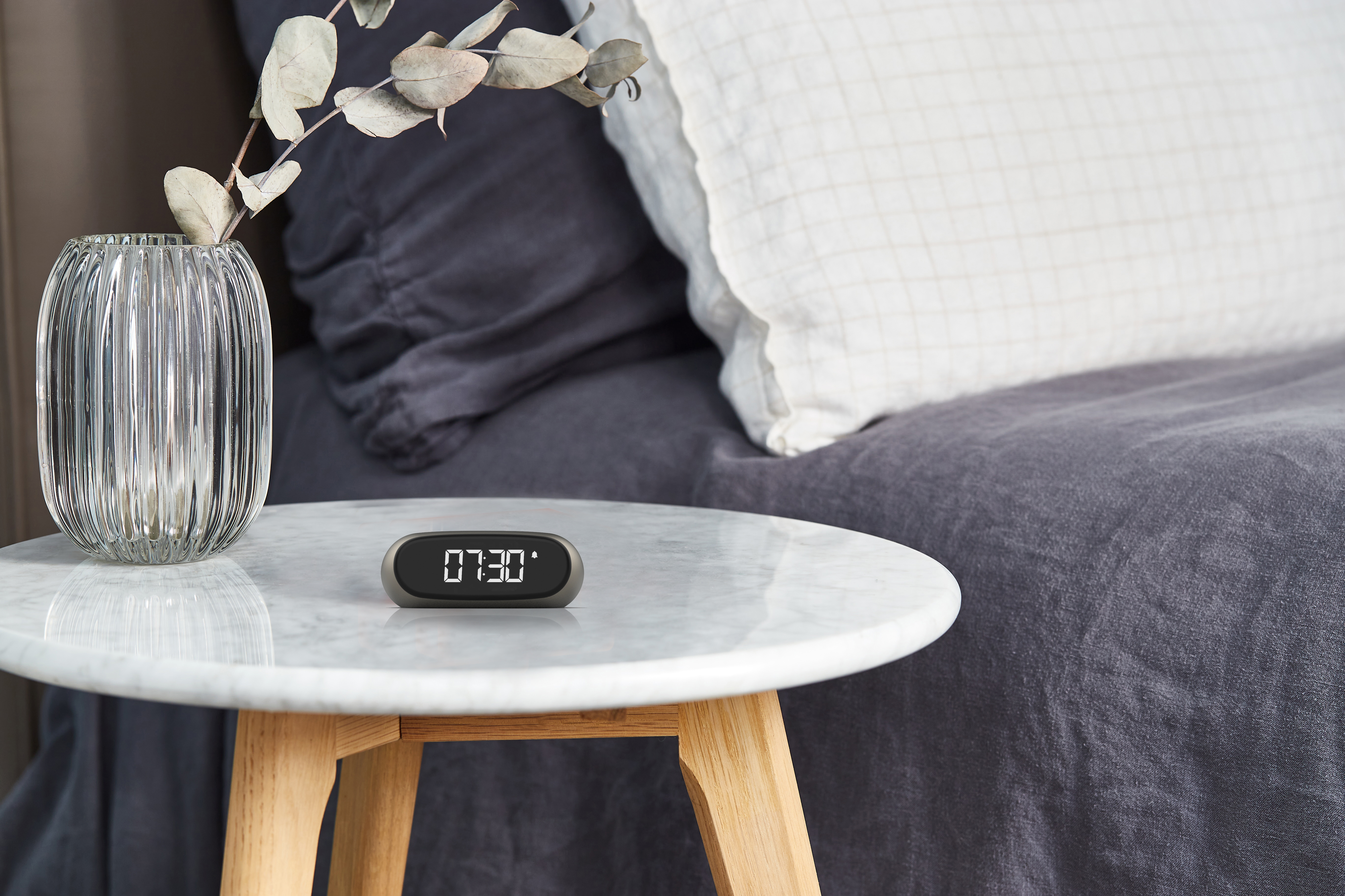 Sveglia su un tavolo, un vaso con una pianta e un letto con cuscini sullo sfondo.