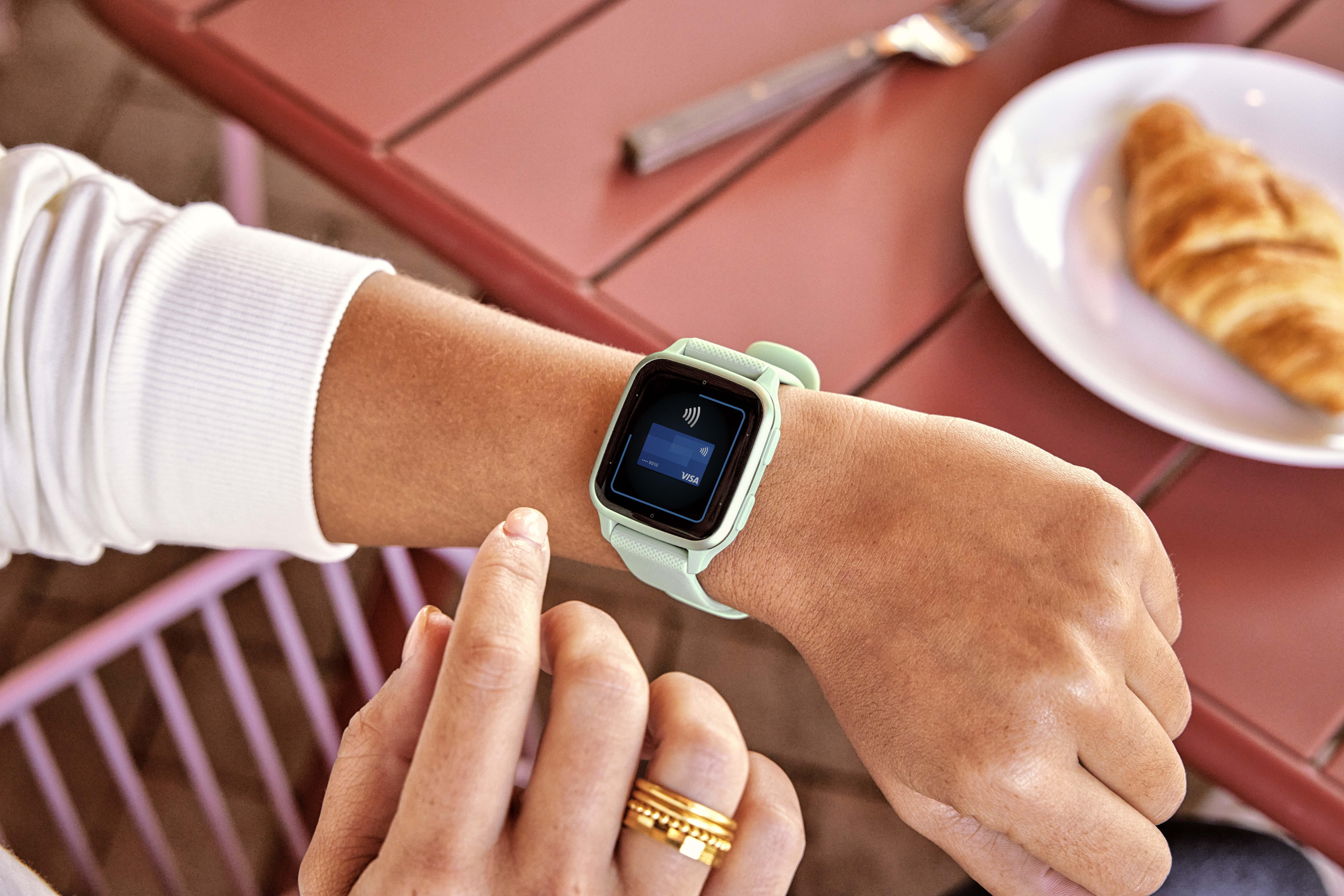 Handgelenk einer Person mit mintfarbener Smartwatch. Es zeigt ein Zahlungssymbol.