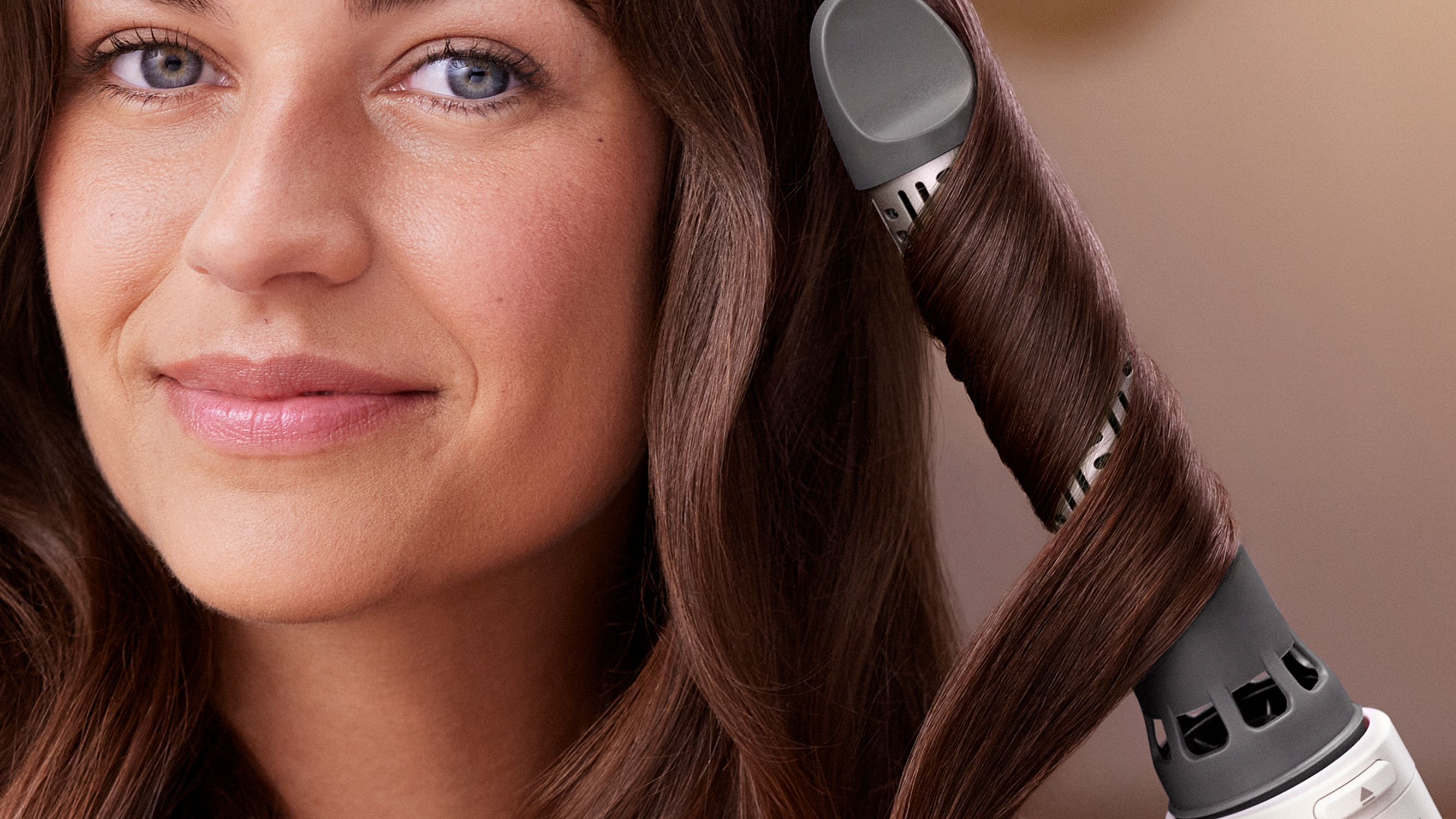 A woman smiles while a curling iron shapes her brown hair into a curl.