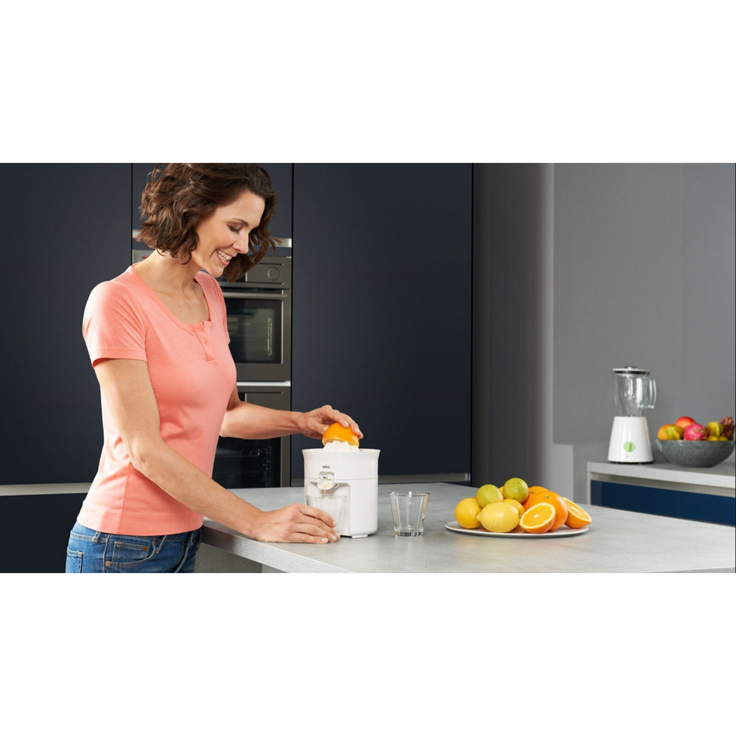 Mujer exprimiendo una naranja con un exprimidor blanco. Plato de frutas y licuadora al fondo.