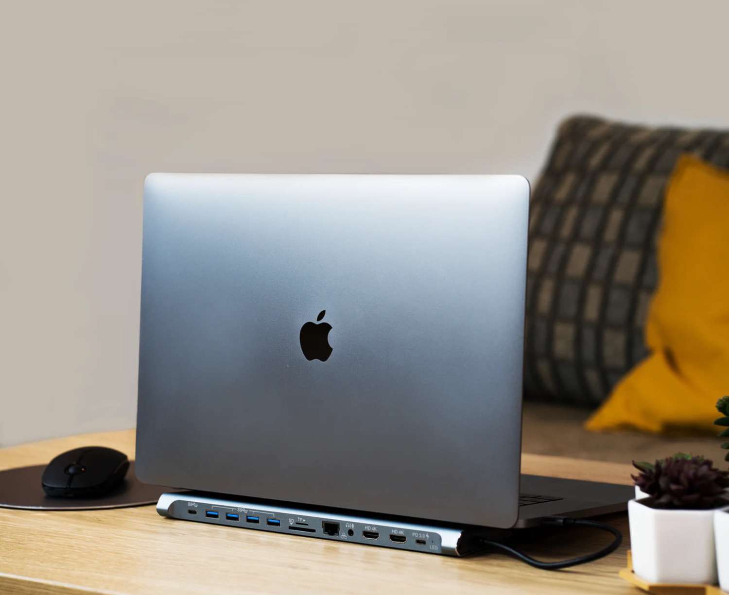 Ordinateur portable sur un bureau connecté à un hub, avec une souris. Un oreiller et une plante en arrière-plan.