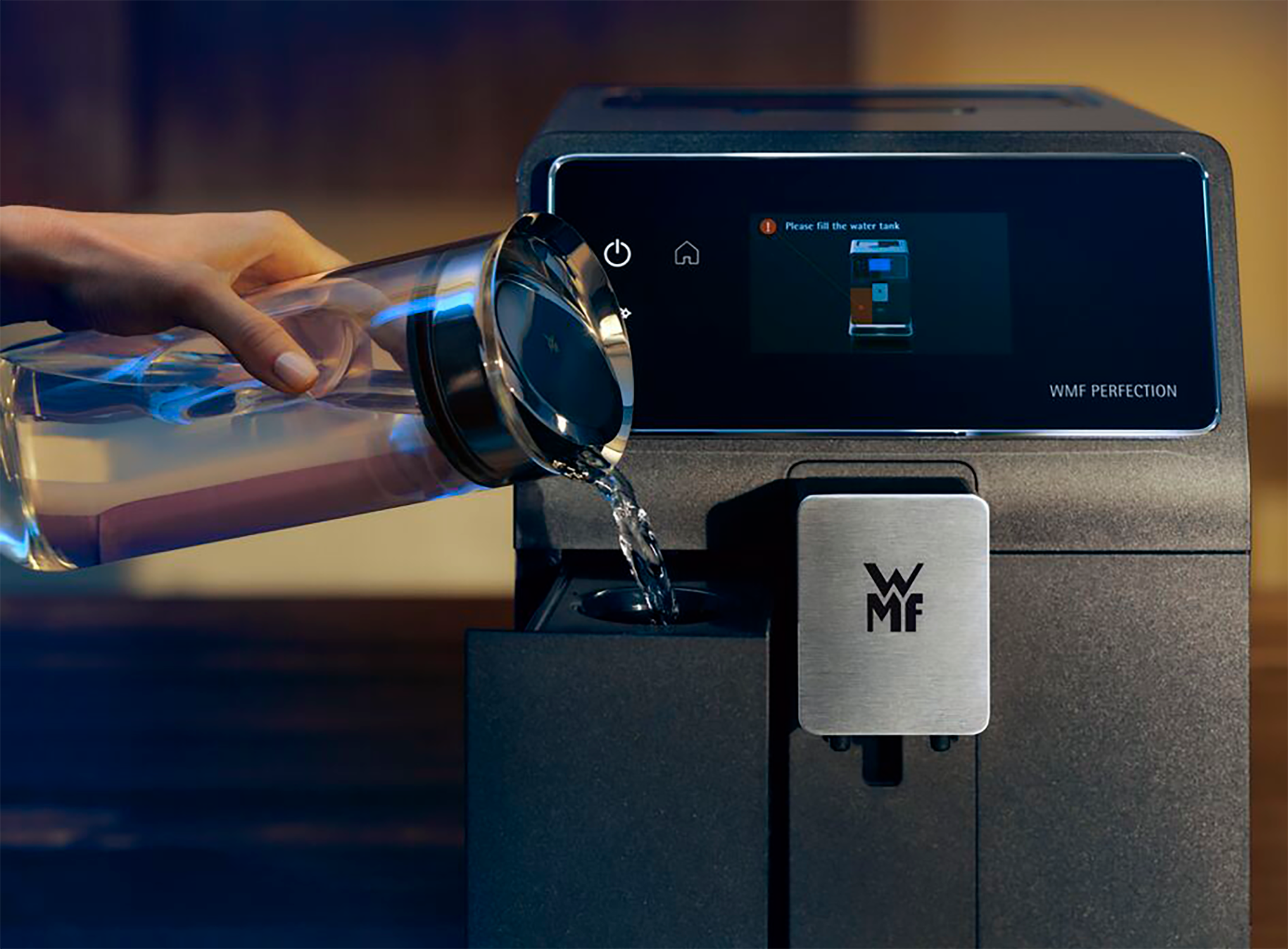 Een hand giet water uit een glazen kan in een zwarte koffiemachine. De machine toont een bericht om water bij te vullen.