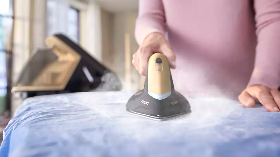 Person ironing a blue shirt. A steam iron is in use. Another appliance is visible in the background.