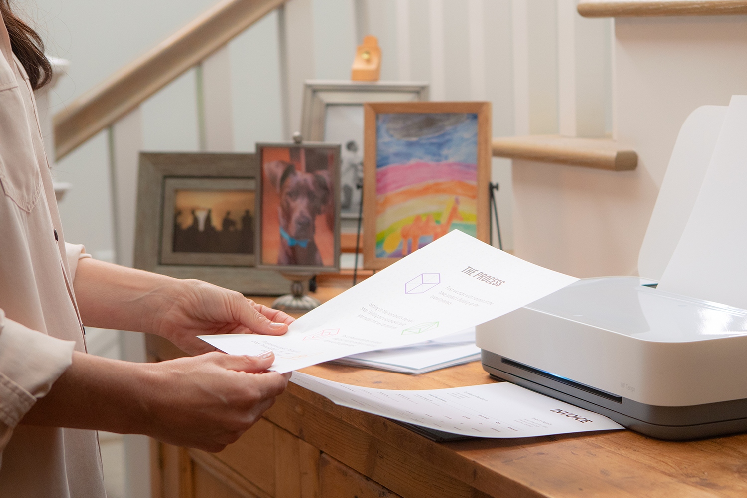 Woman's hands hold papers near a printer, framed photos in background.
