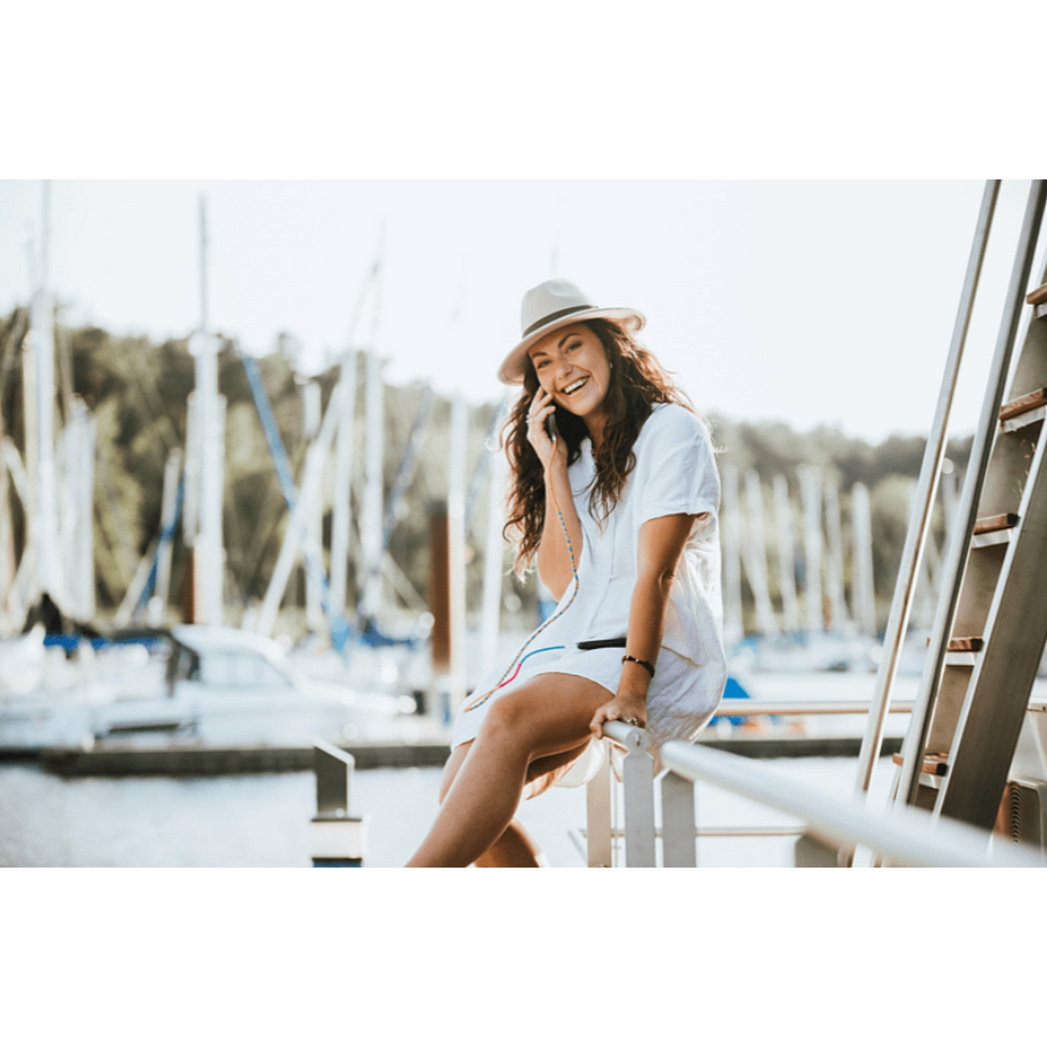 Mujer con vestido blanco y sombrero sentada en un muelle, hablando por teléfono, con barcos de fondo.