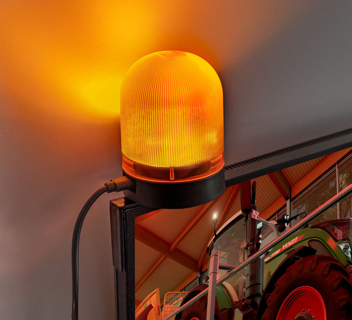 Ein orangefarbenes Warnlicht an einer schwarzen Halterung. Ein Traktor ist im Hintergrund.