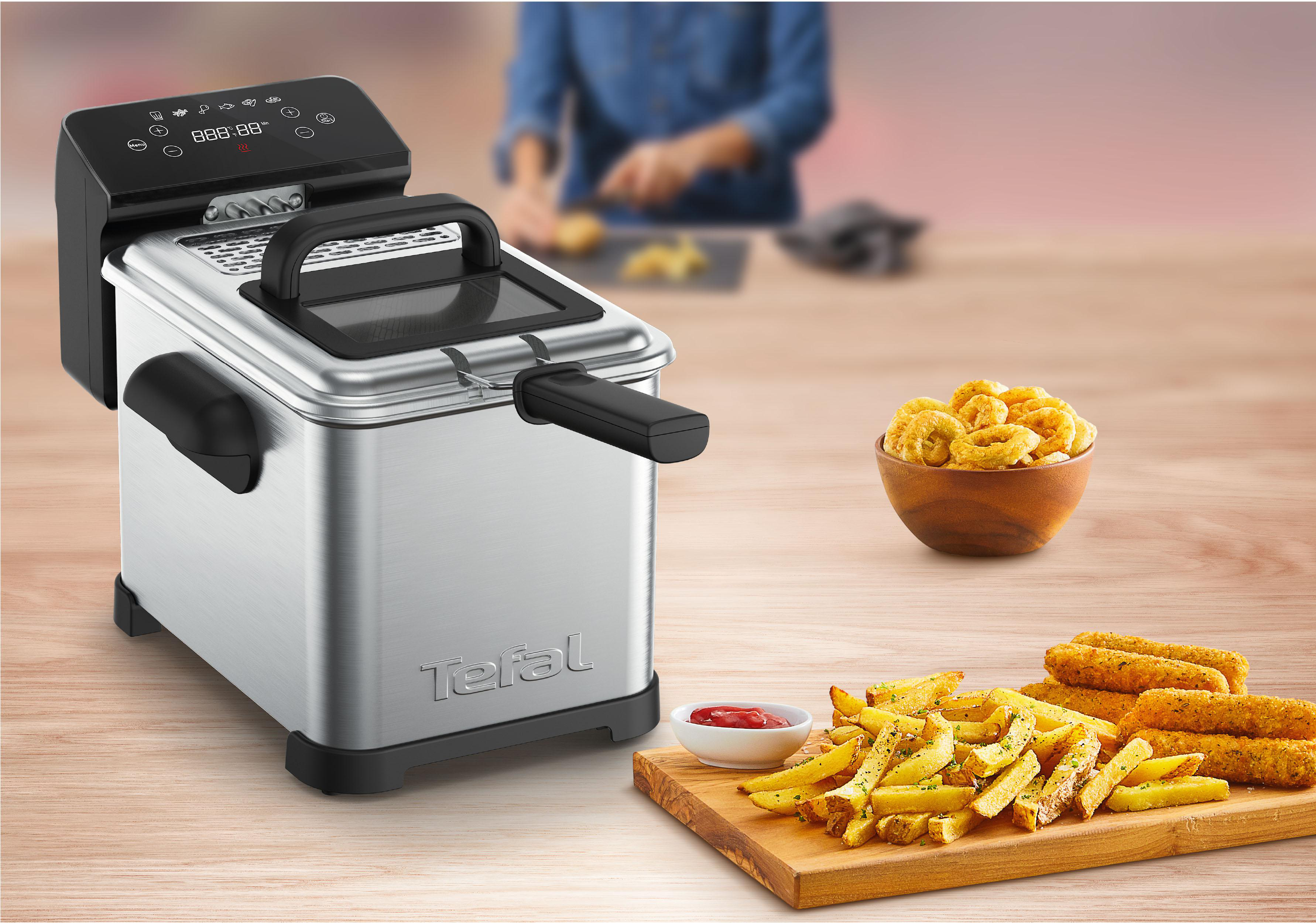 Silberne Tefal Fritteuse mit Pommes Frites, Mozzarella-Sticks und Zwiebelringen auf dem Tisch.