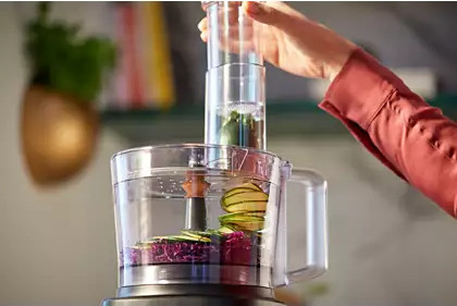 A food processor is being used with vegetables inside and a hand adding to it.