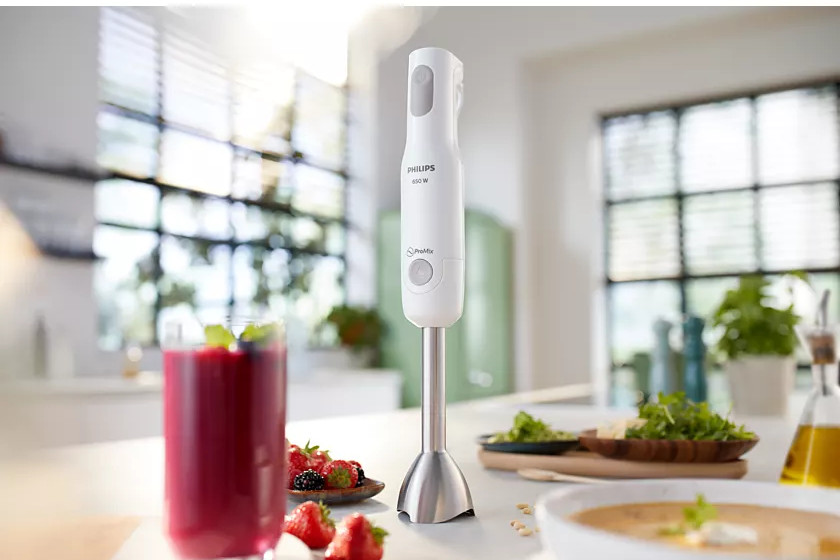 A white hand blender stands on a counter, with a glass of smoothie and ingredients nearby.