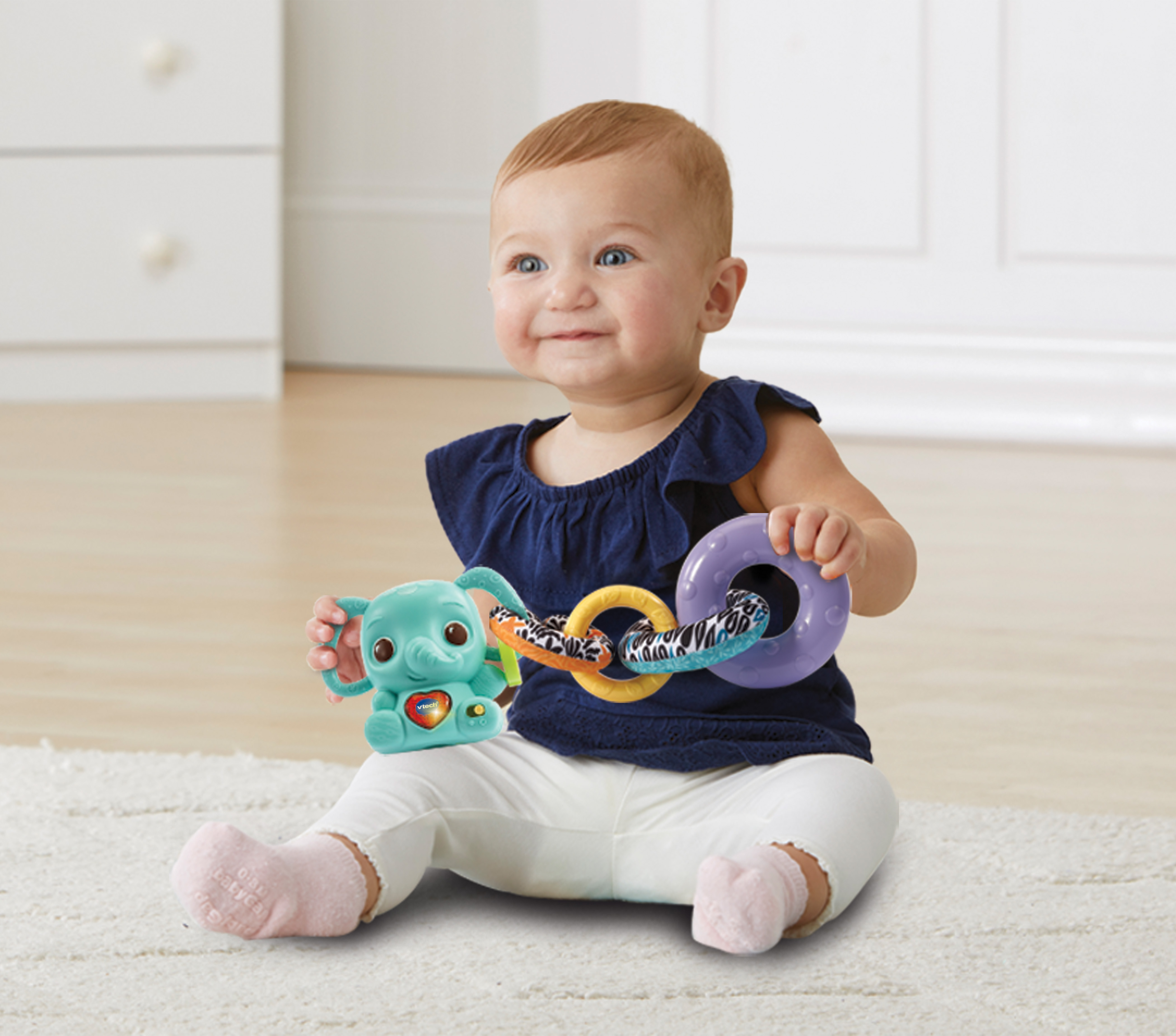 Ein Baby sitzt und spielt mit einem blauen Elefantenspielzeug und bunten Ringen, lächelnd.
