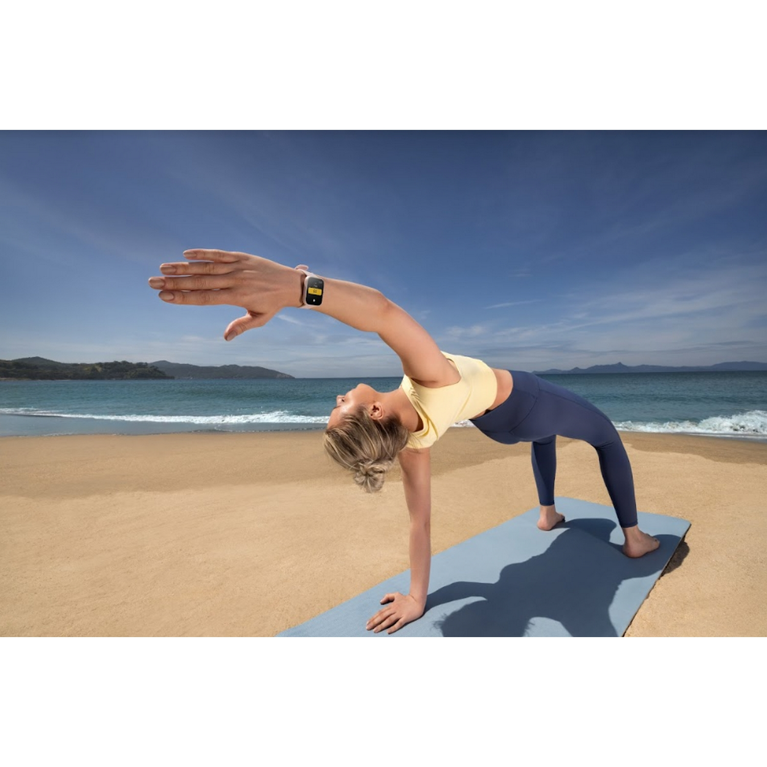 Mujer haciendo yoga en una playa. Lleva top amarillo, pantalones azules y un smartwatch en la muñeca.