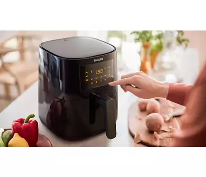 Black air fryer with digital display, a hand touching it, vegetables, and kitchen background.