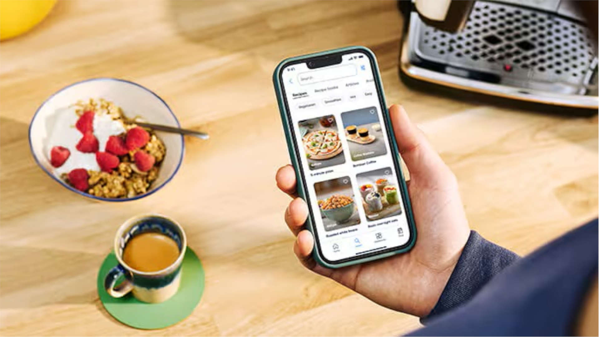 Personne tenant un téléphone avec une application, petit-déjeuner et café à proximité.