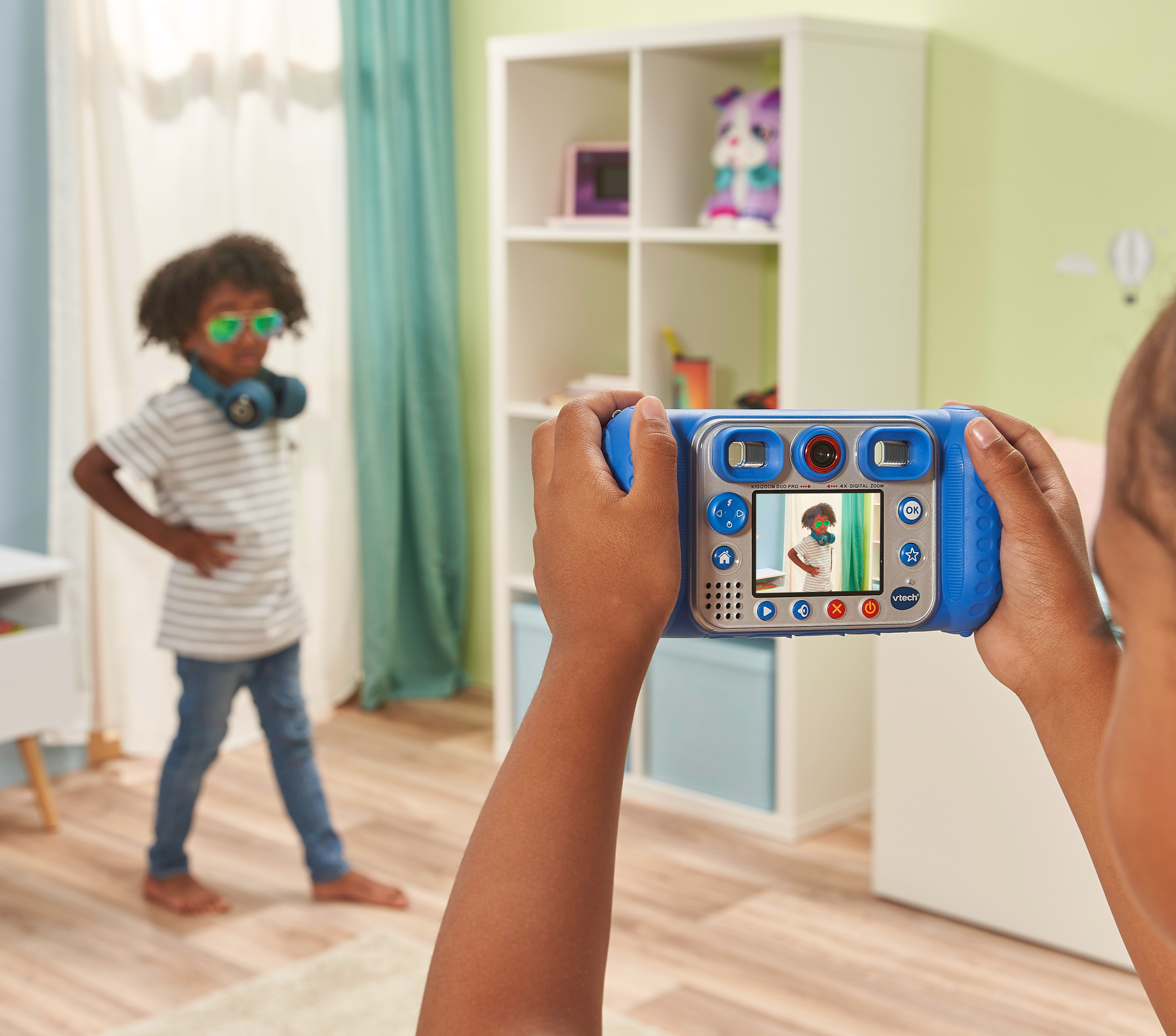 Ein Kind macht mit einer blauen Spielzeugkamera ein Foto von einem anderen Kind in einem Raum.