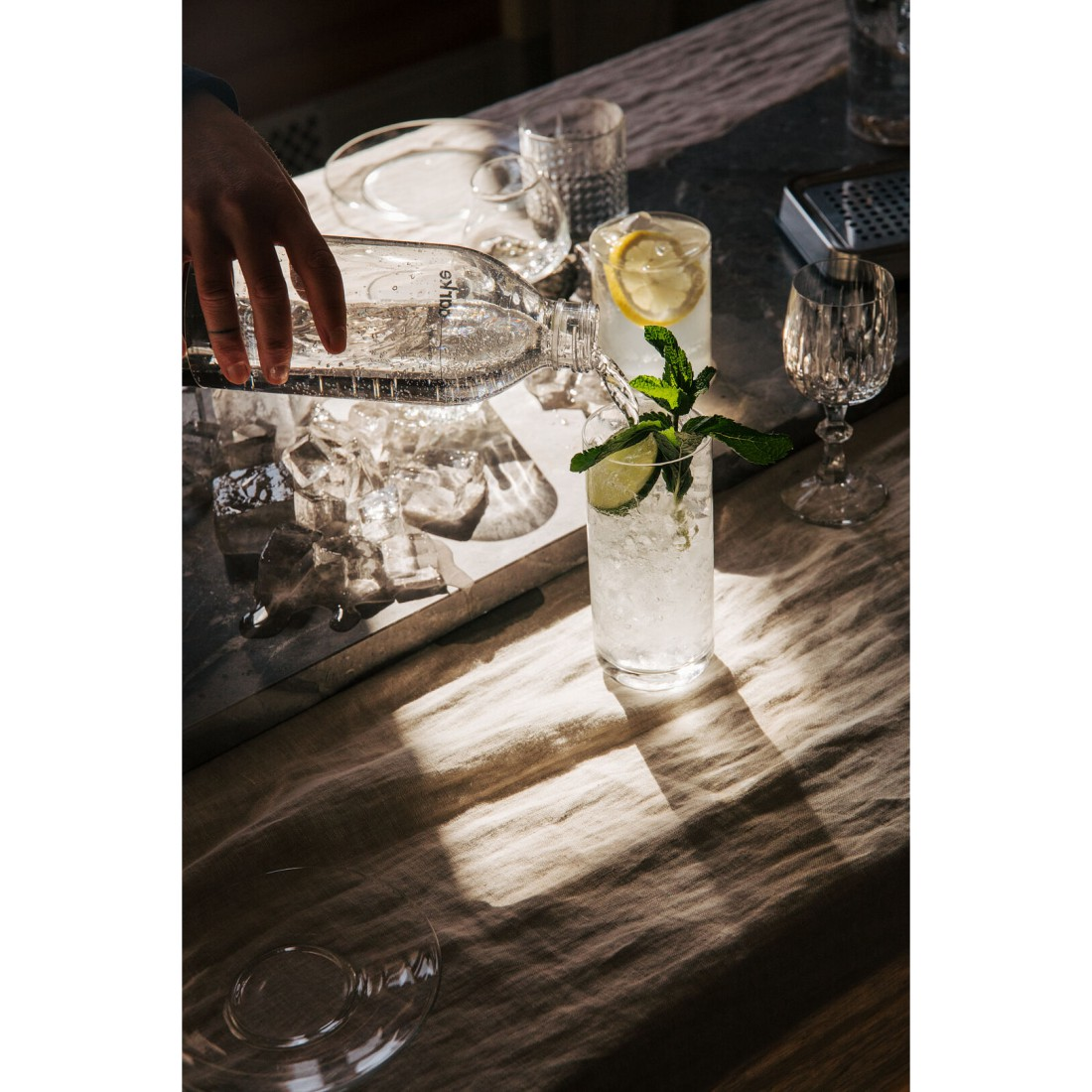 Eine Hand, die Wasser aus einer Flasche in ein Glas mit Eis, Zitrone, Minze und Limette gießt.