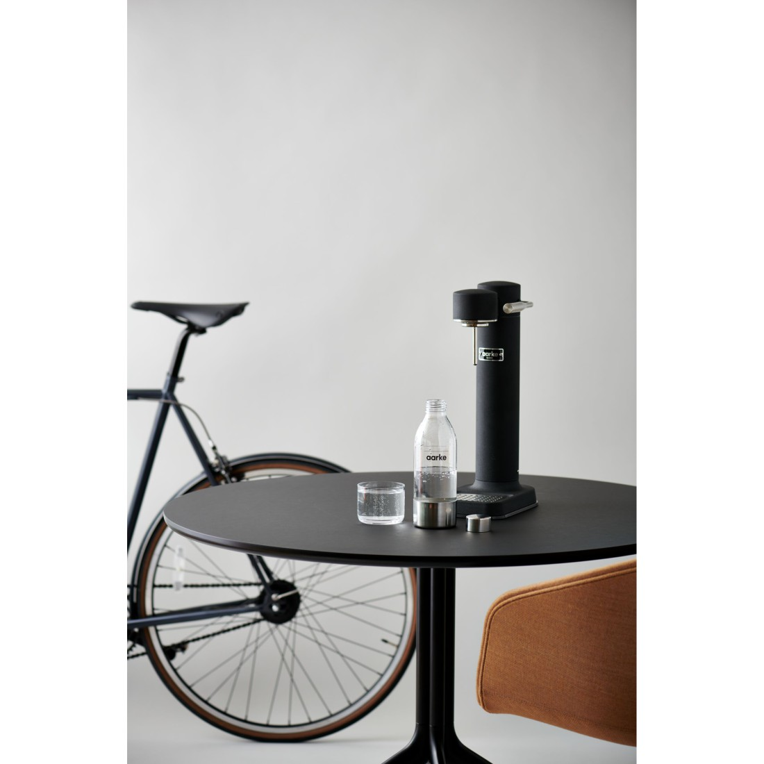Ein schwarzer Sodabereiter, Glas und Flasche auf einem Tisch. Im Hintergrund ein Fahrrad.