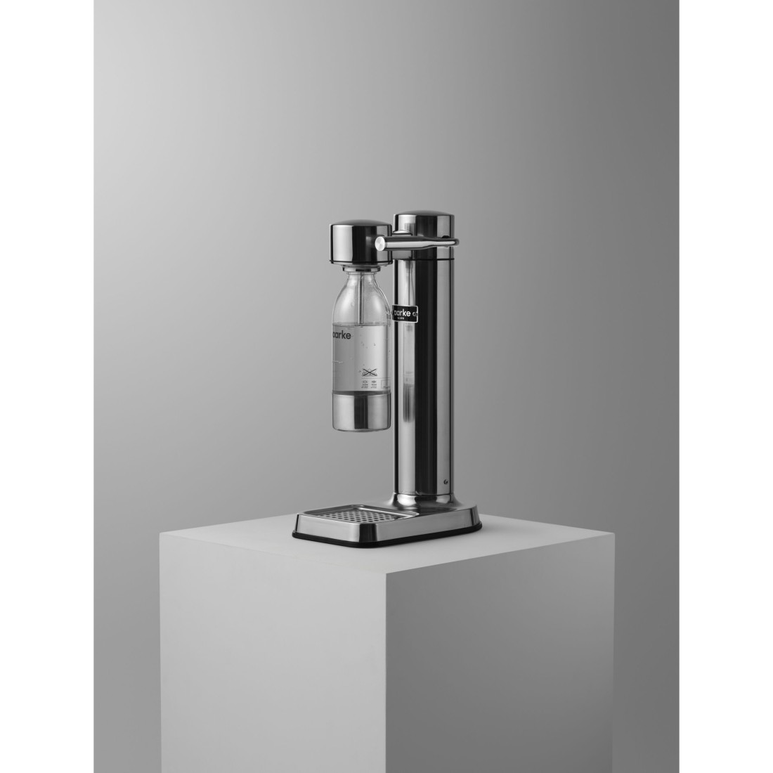 Soda-Maker aus Edelstahl auf weißem Block, Glasflasche befestigt, grauer Hintergrund.