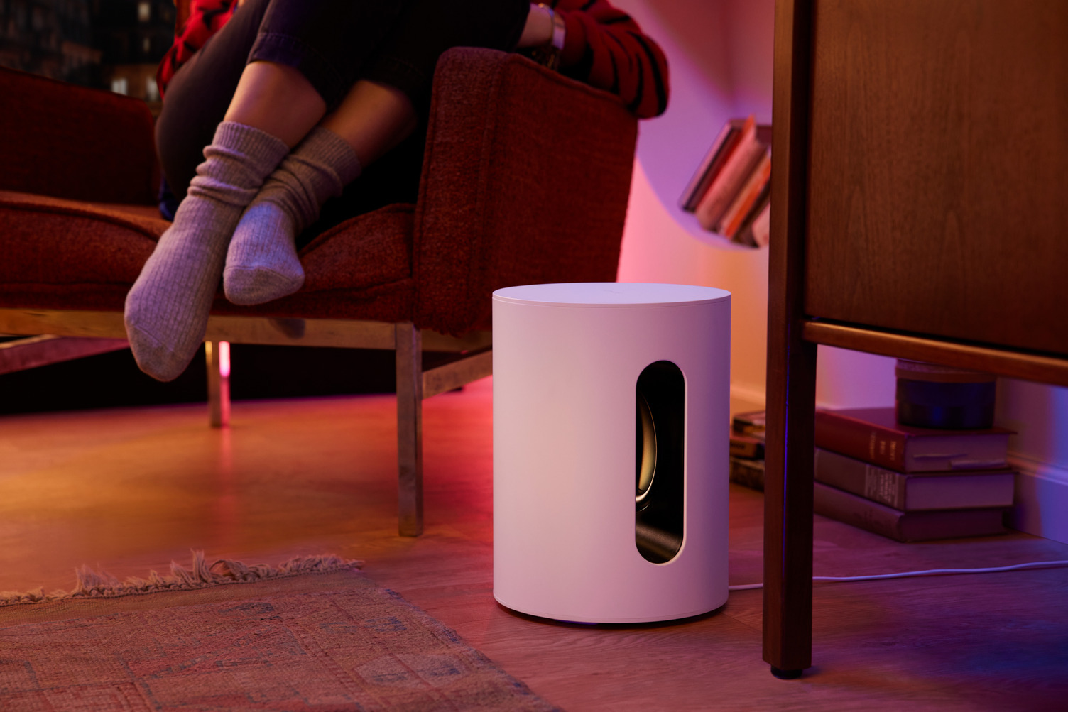 A white subwoofer with a curved cutout and a person in socks sitting on a red couch in the background.