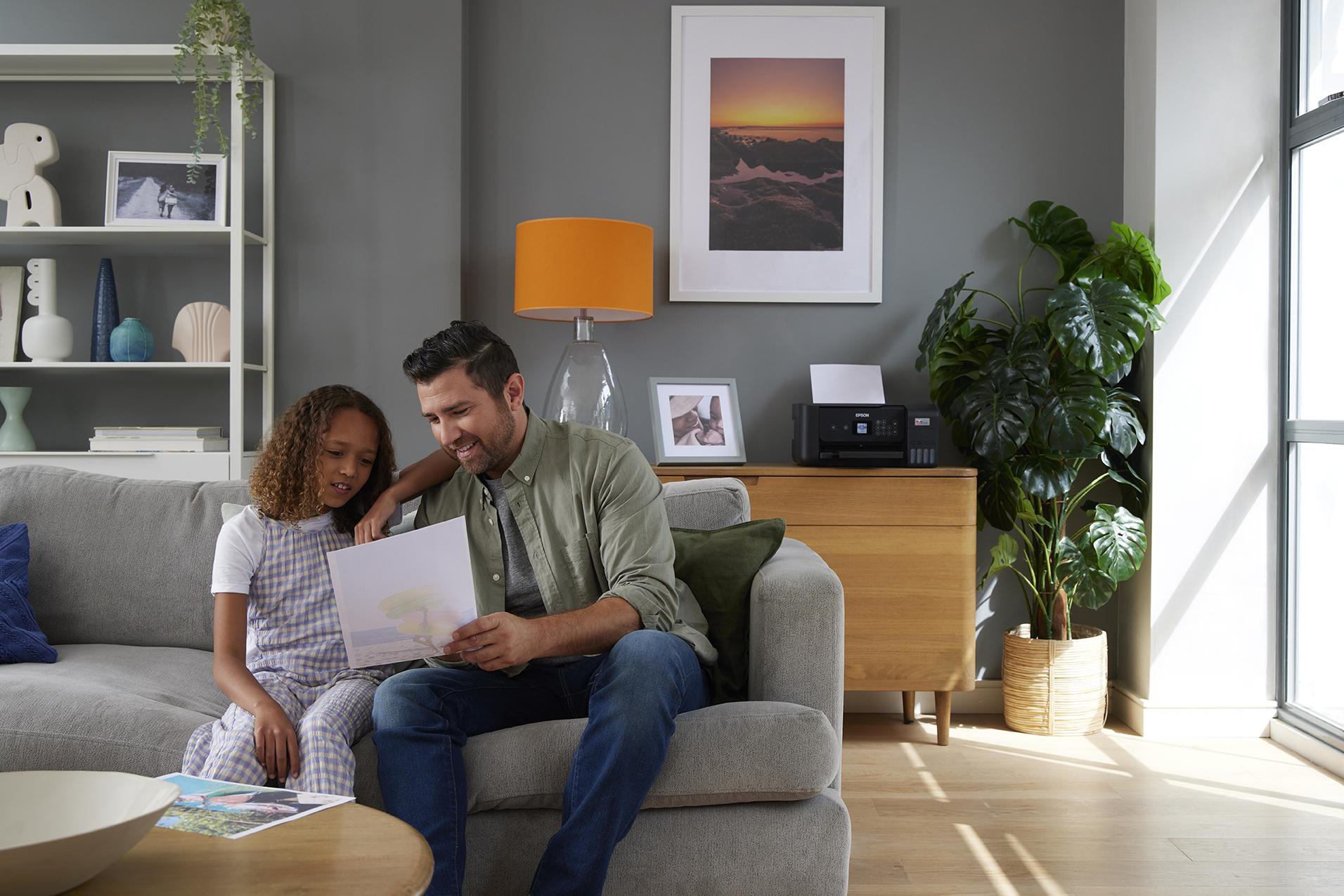 Ein Vater und eine Tochter sitzen auf einem Sofa und betrachten Fotos. Ein Drucker befindet sich rechts.
