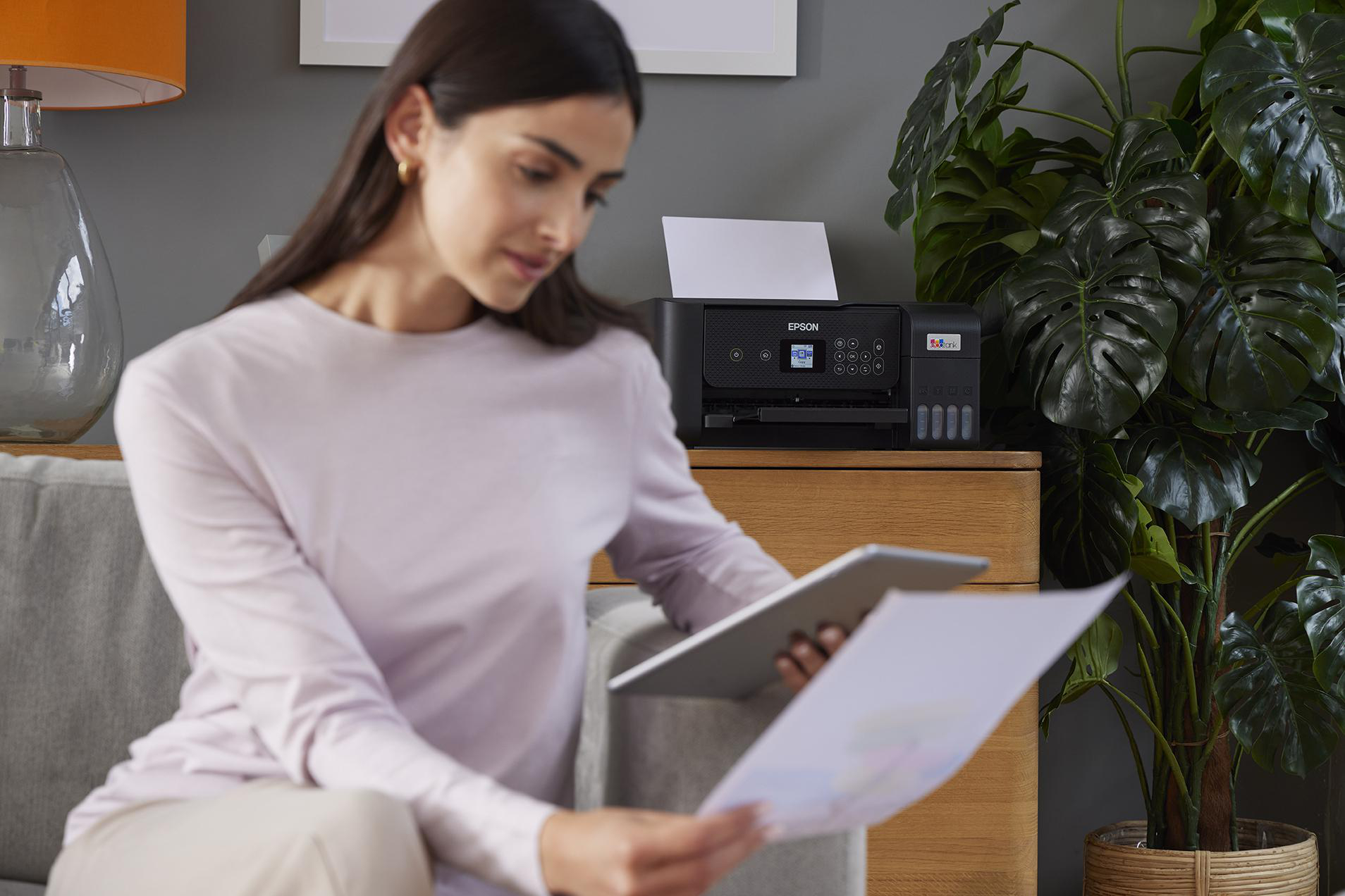 Frau sitzt auf Couch, schaut auf Tablet, Drucker und Pflanze.