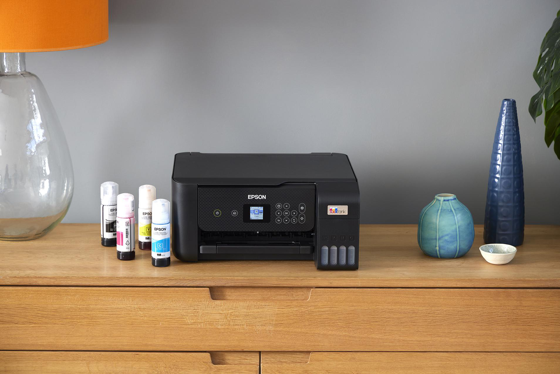 Schwarzer Epson-Drucker auf einem Holztisch mit Tintenpatronen. Dekorative Gegenstände im Hintergrund.