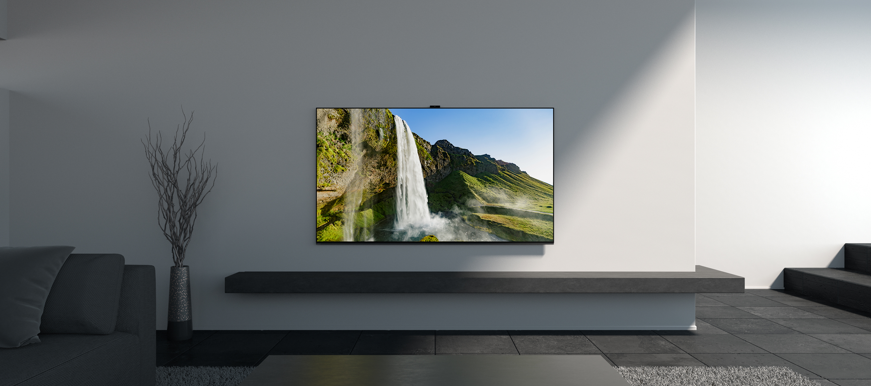 Ein großer Fernseher an einer weißen Wand, der eine Wasserfallszene zeigt. Ein graues Regal darunter, mit Sofa.
