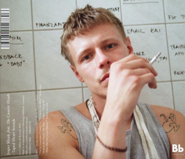 Ein Mann mit Tattoos raucht eine Zigarette und blickt in die Kamera. Text an der Wand.