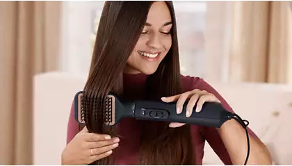 A woman using a hair straightening brush, smiling, with long brown hair.