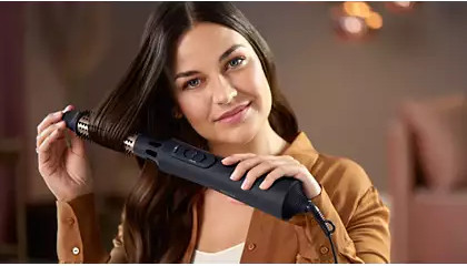 A woman uses a black hair dryer, styling her hair, with a soft background.