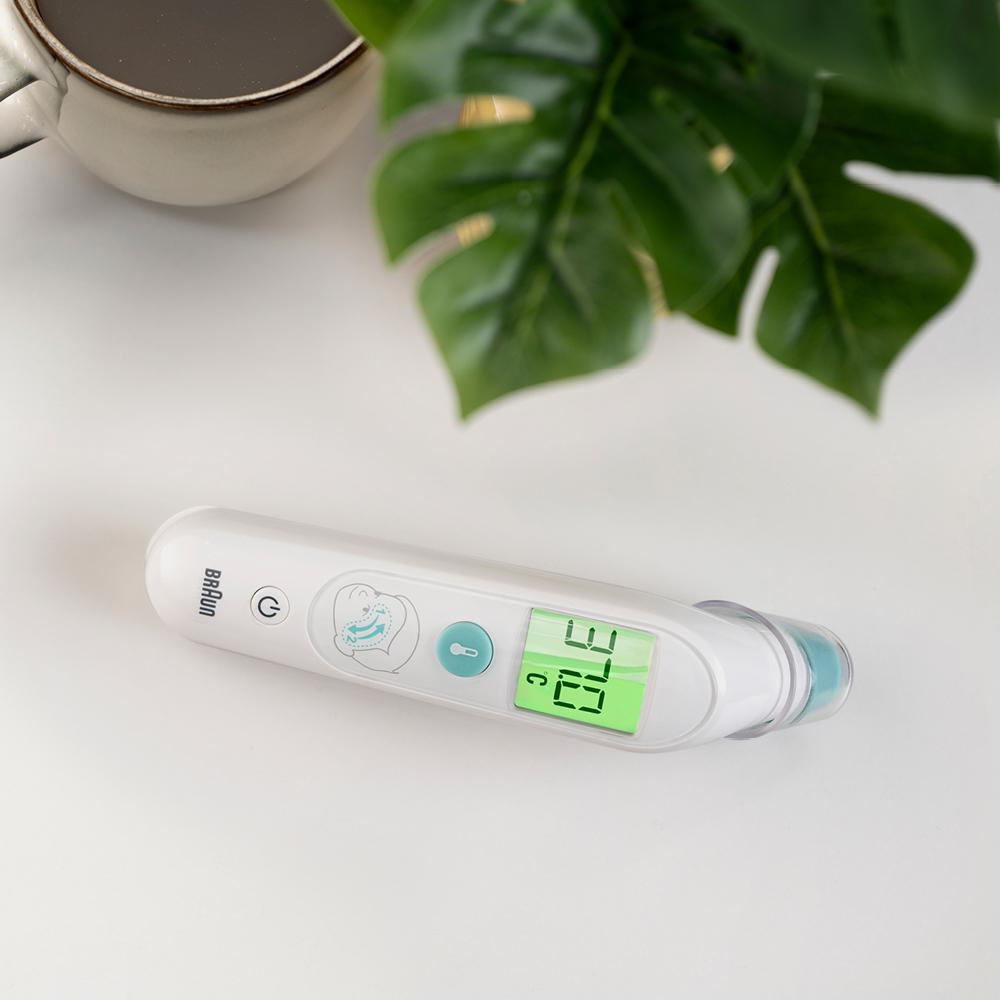 Weißes Braun-Thermometer, zeigt 37,0°C an, neben einer Tasse und einer Pflanze auf weißer Oberfläche.