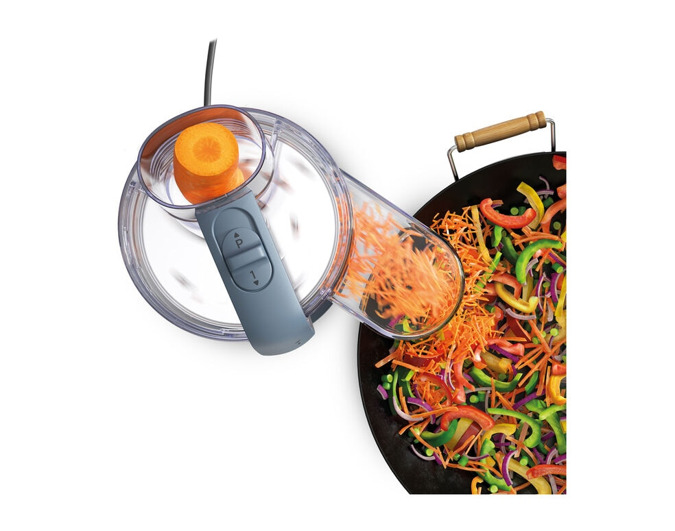 A food processor slicing a carrot into a pan of vegetables.