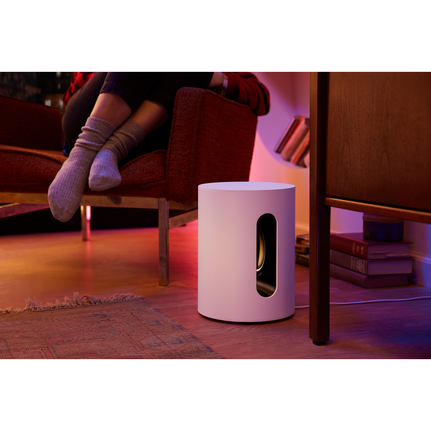Een persoon zit in een rode fauteuil. Een witte subwoofer staat ernaast.