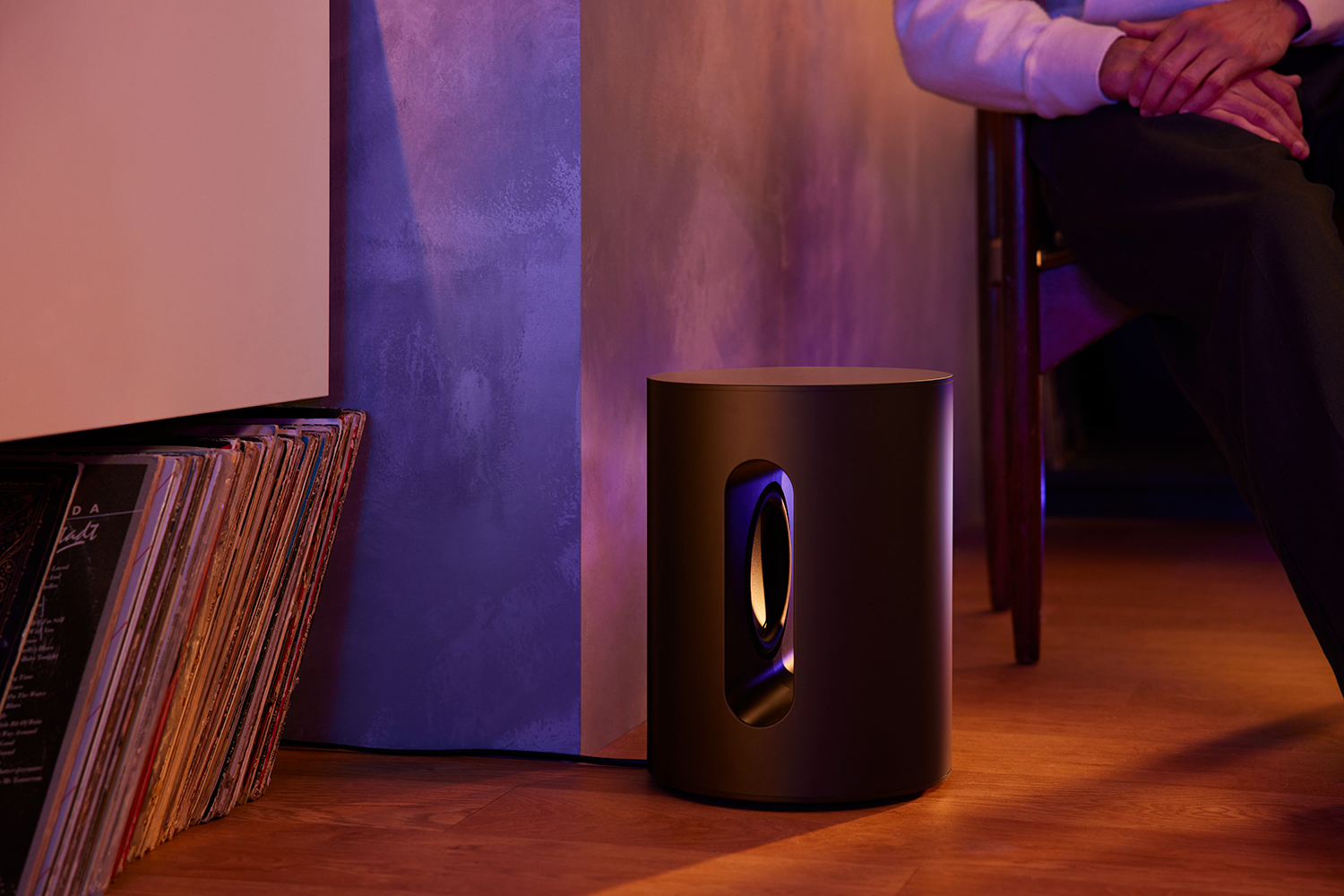 Zwarte subwoofer bij een muur, met een zittende persoon. Platen links.