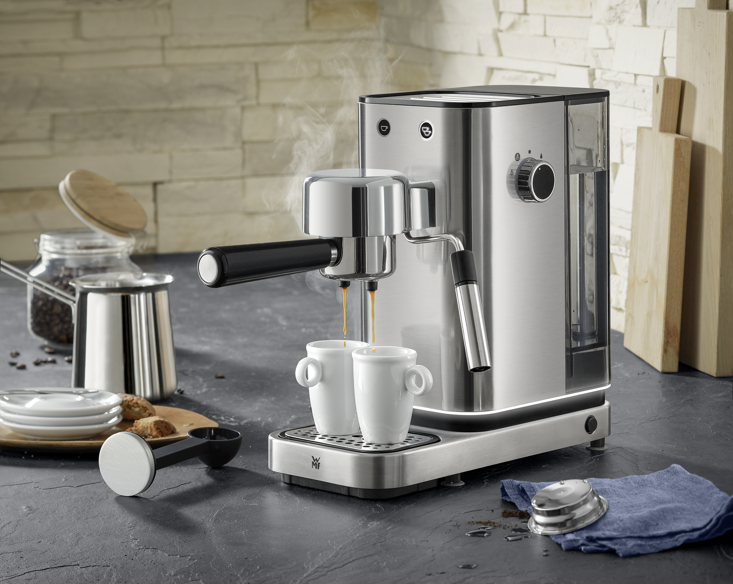 Máquina de espresso con tazas, café y accesorios. El vapor se eleva. Fondo: cocina.