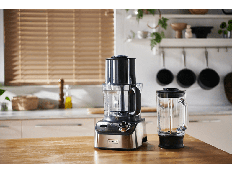 Un robot culinaire et un blender sur un comptoir en bois. Une fenêtre avec des stores et des ustensiles de cuisine en arrière-plan.