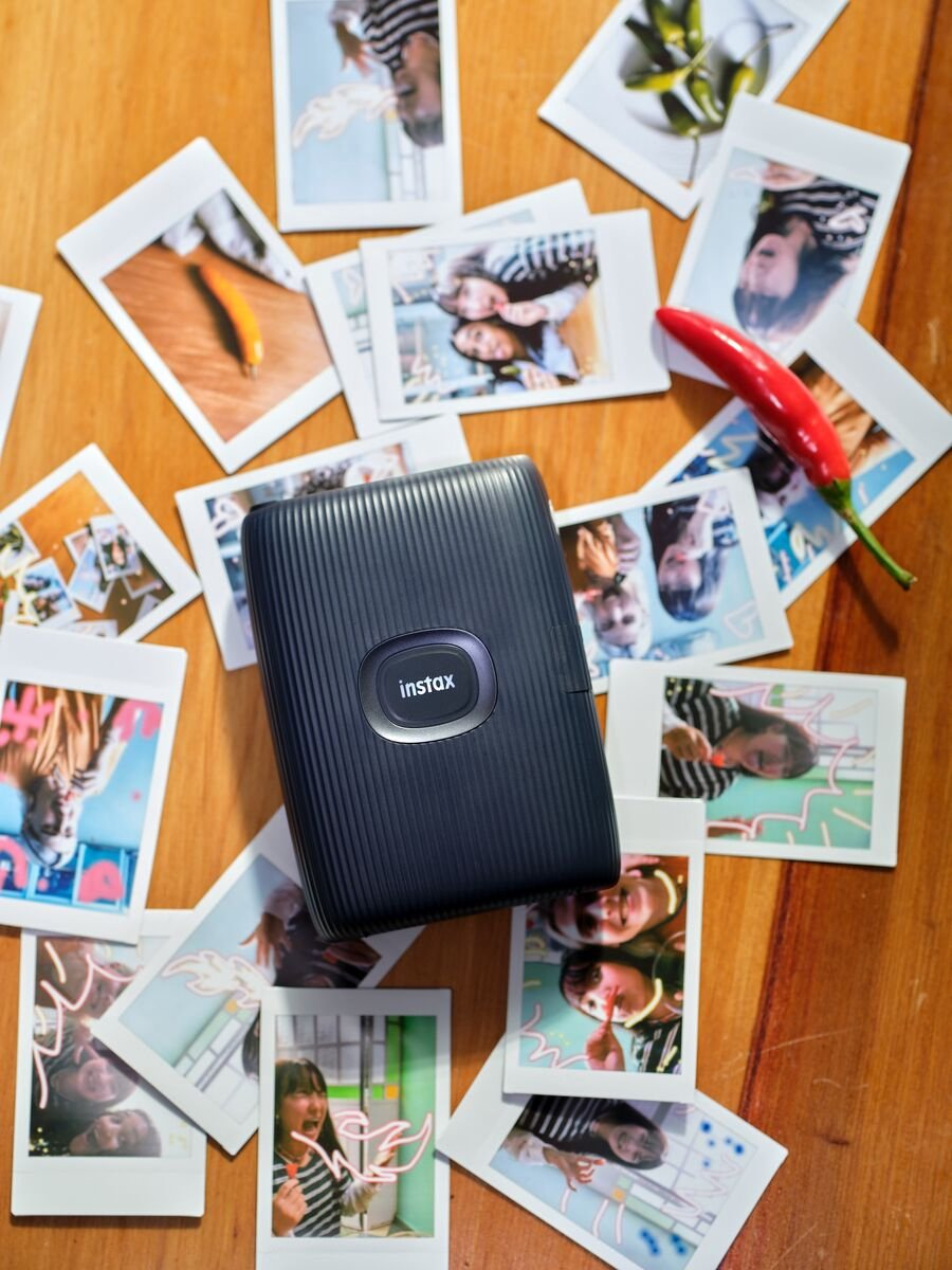 Une imprimante Instax noire entourée de photos et d'un piment rouge.