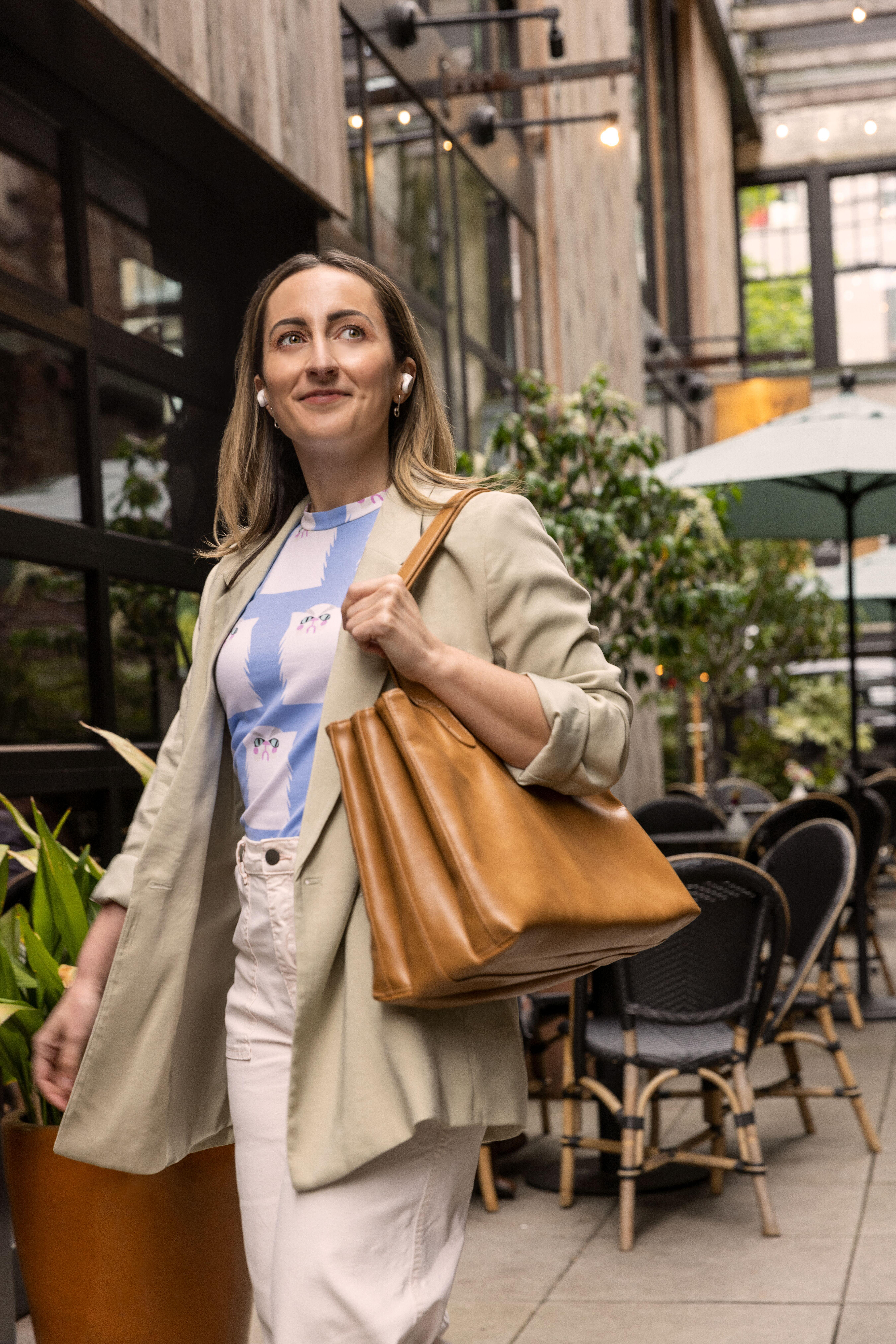 Eine Frau mit beigem Blazer und Tasche geht vor einem Café. Sie trägt Ohrhörer und lächelt.