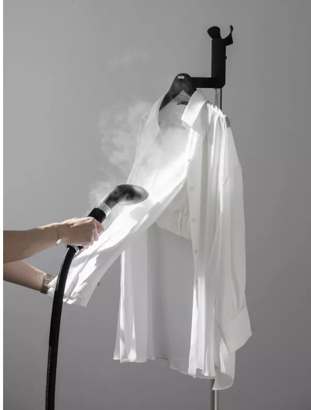 Une main repasse à la vapeur une chemise blanche sur un cintre, la vapeur monte. Le fond est un mur gris clair.