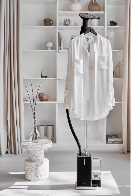 Chemise blanche suspendue sur un défroisseur, dans une pièce blanche avec étagères et déco.