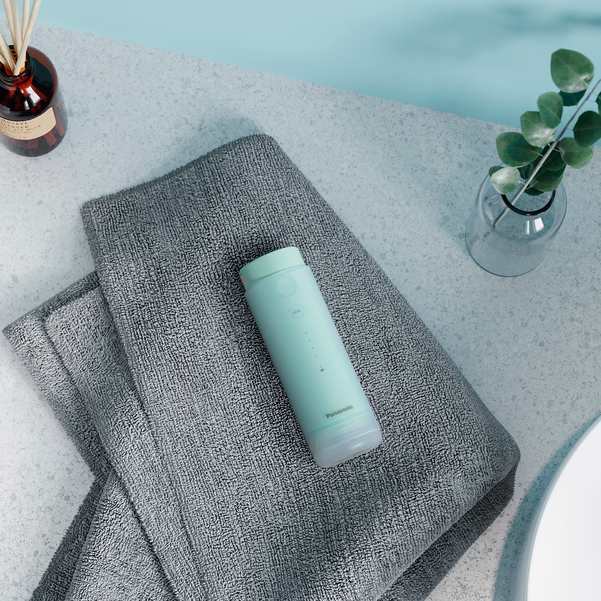 Une bouteille d'eau vert clair sur des serviettes grises avec une petite plante et un flacon de parfum.