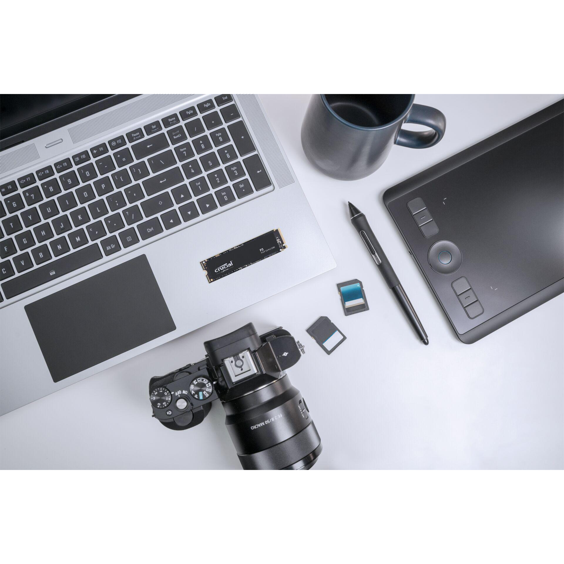 Draufsicht mit Laptop, Kamera, SSD, SD-Karten, Stift und Becher auf weißer Oberfläche.