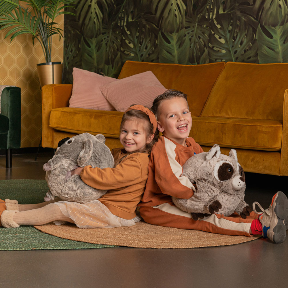 Zwei Kinder mit Plüschtieren sitzen auf einem Teppich vor einem gelben Sofa.