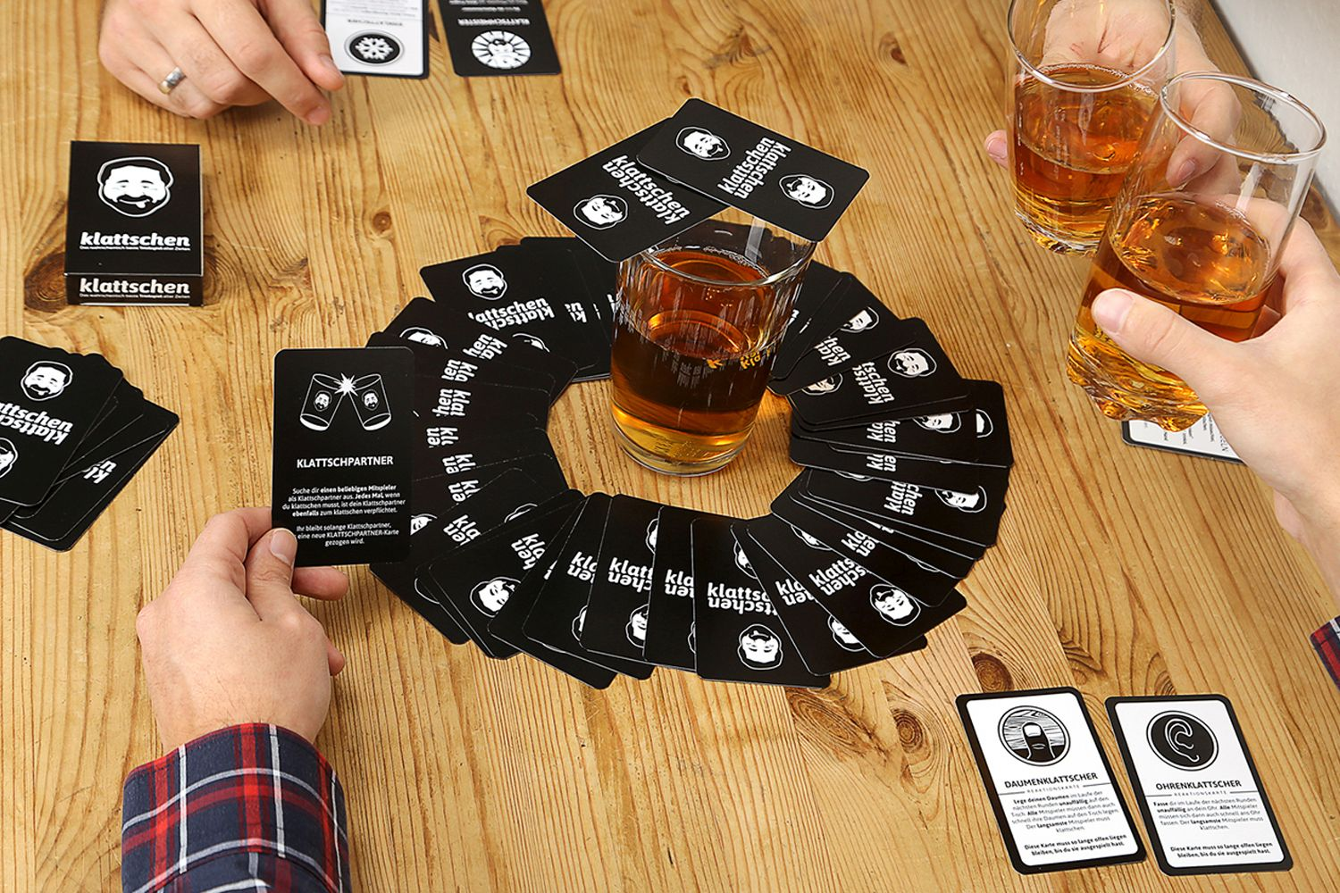 Leute spielen ein Kartenspiel, umgeben von Karten. Zwei Leute halten Gläser mit Flüssigkeit, das Spiel ist auf einem Holztisch.