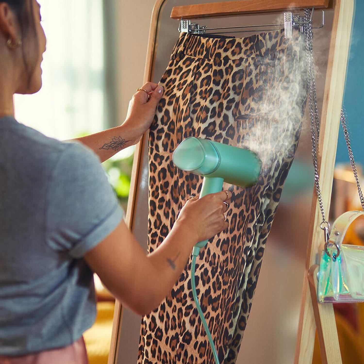Donna che stira pantaloni leopardati su un attaccapanni con un vaporizzatore verde.