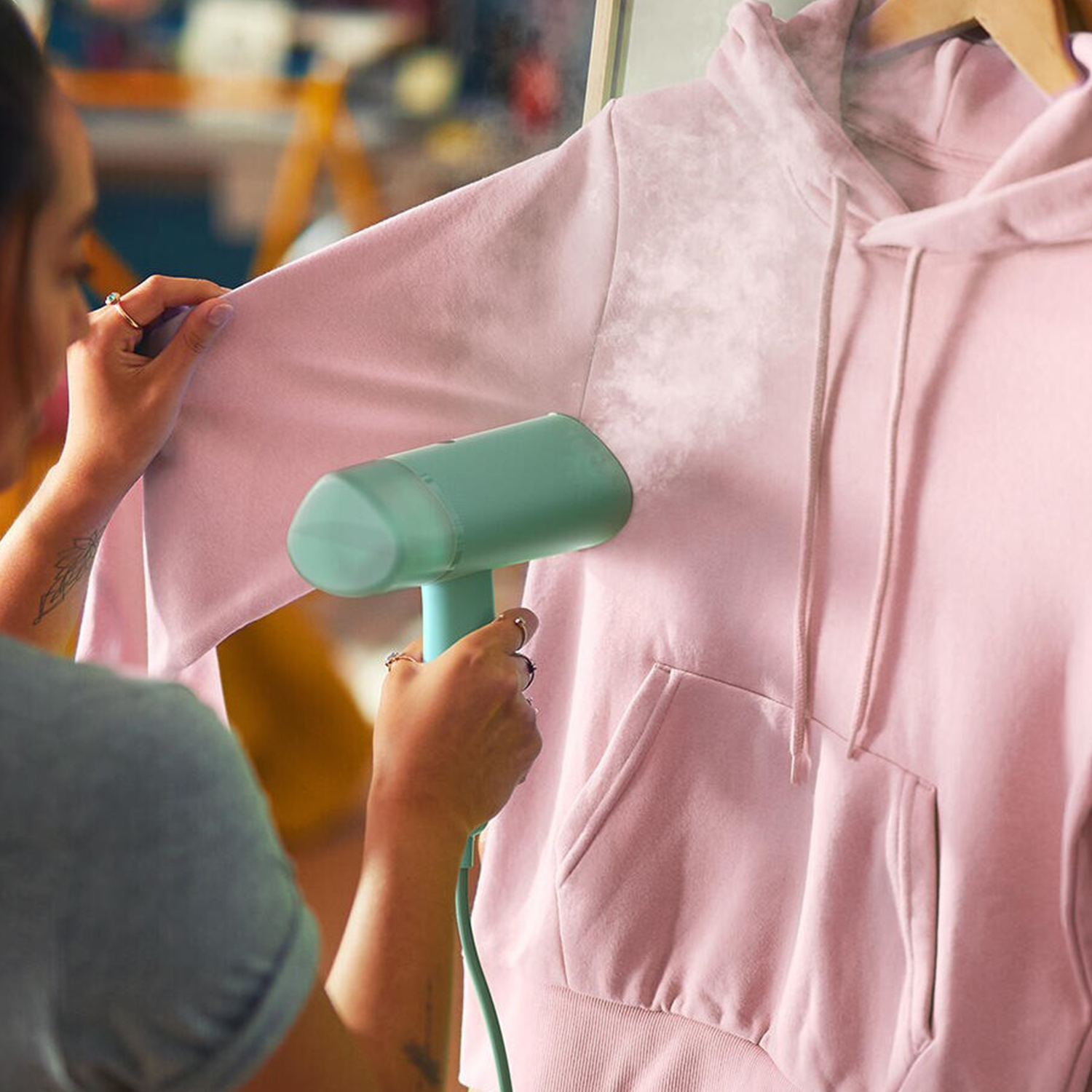 Una persona che stira una felpa con cappuccio rosa con un vaporizzatore portatile, primo piano.
