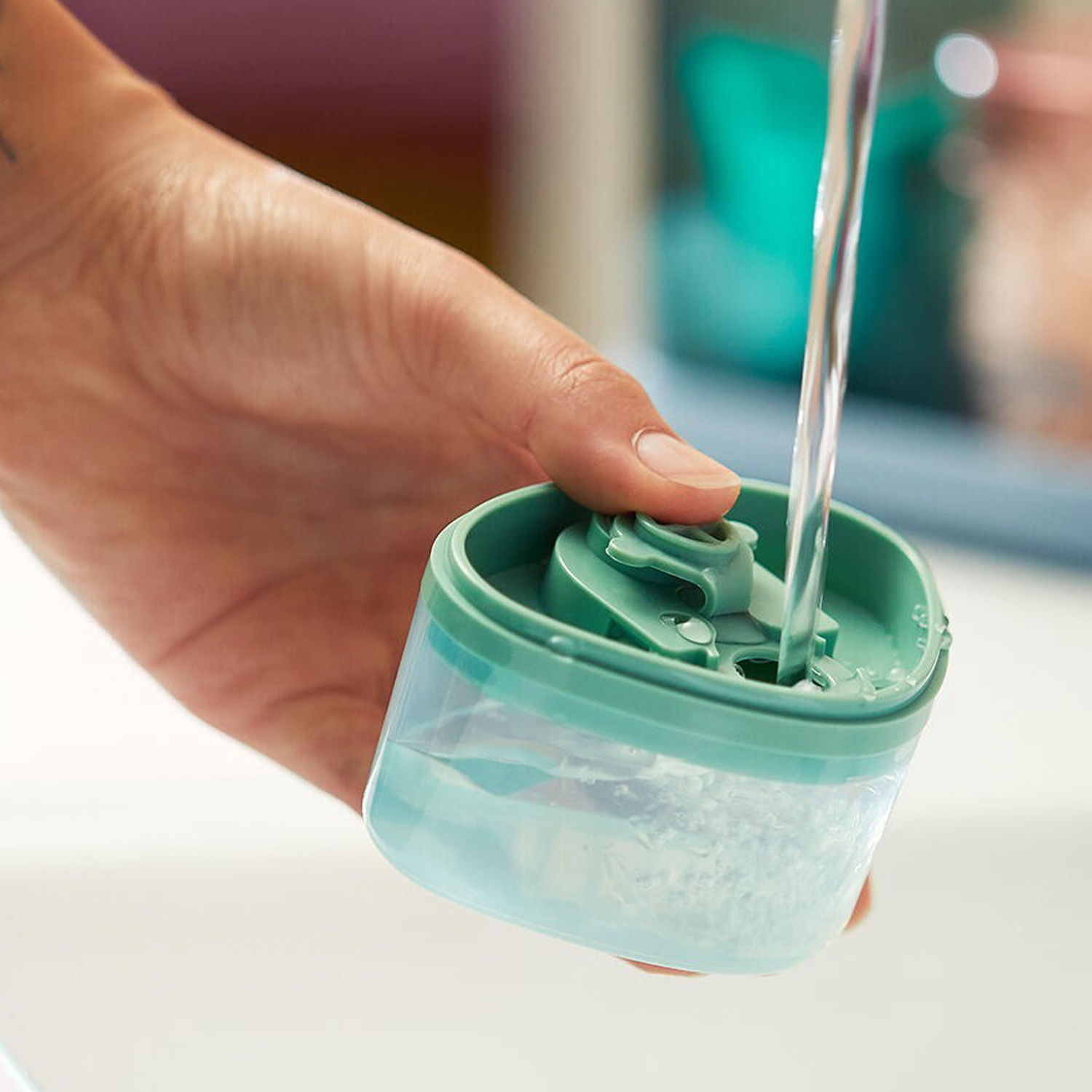 Una mano versa acqua in un contenitore verde acqua e trasparente su un lavandino bianco.