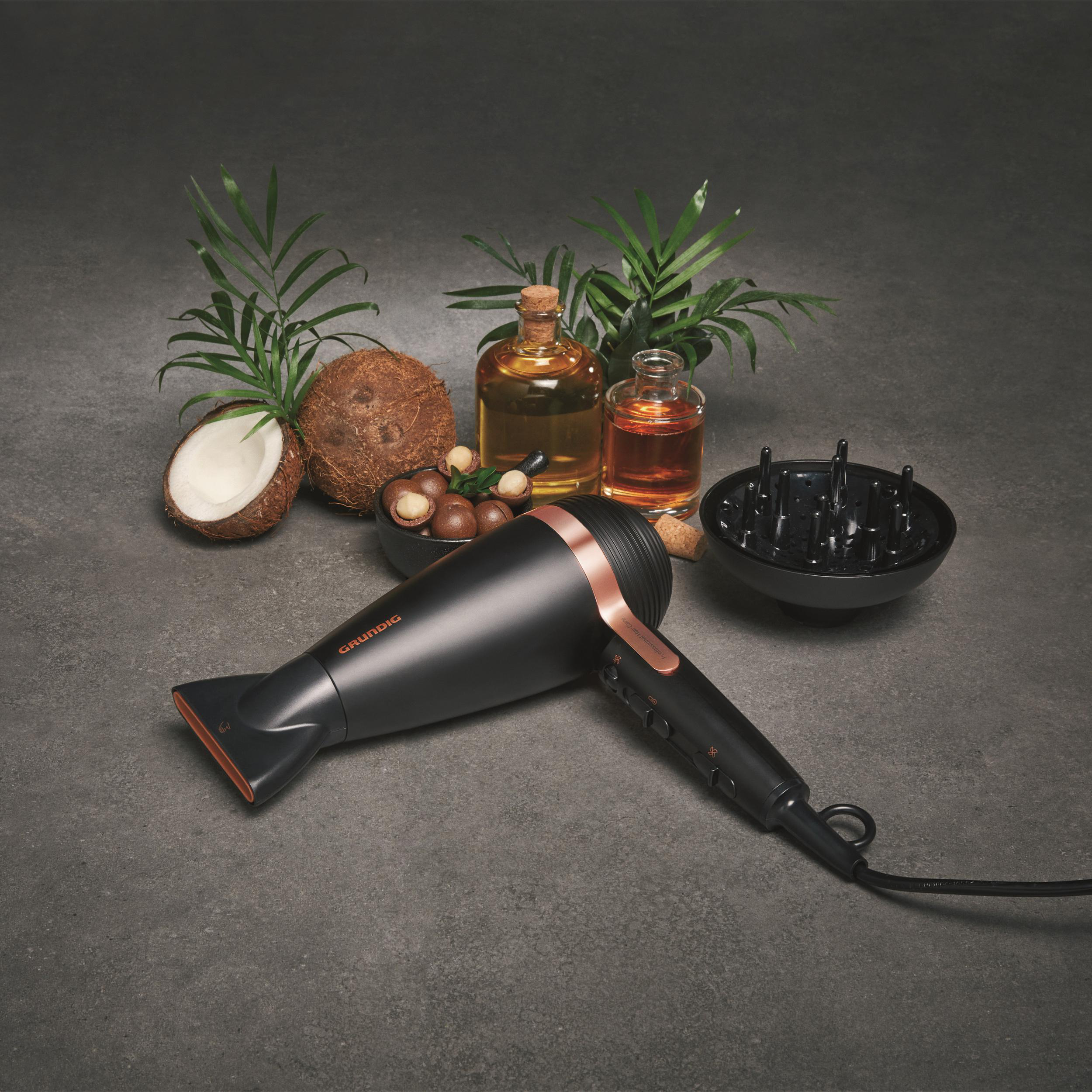 Ein Haartrockner, Kokosnuss, Öle und ein Diffusor liegen auf grauer Oberfläche mit grünen Pflanzen.